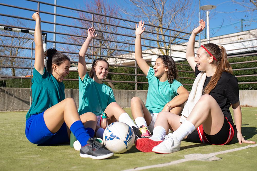Portrait of happy girls talking on football field. Four pretty Caucasian girls in sportswear sitting on ground, raising hands to demonstrate belief in victory. Healthy lifestyle and team sport concept